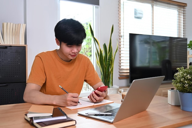 Feliz homem asiático segurando o telefone inteligente e fazendo anotações no caderno.