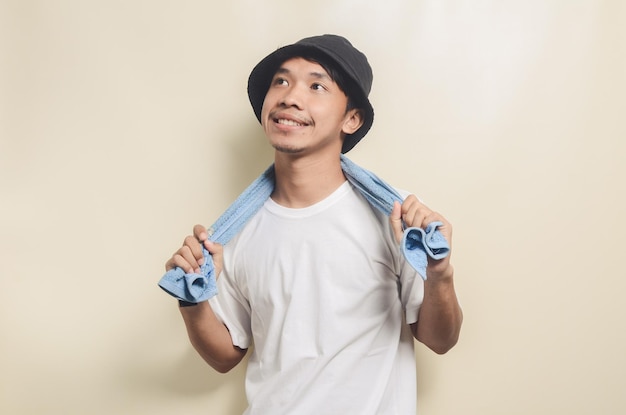 Feliz homem asiático vestindo camiseta branca brilhante com chapéu preto carregando toalha azul em fundo isolado