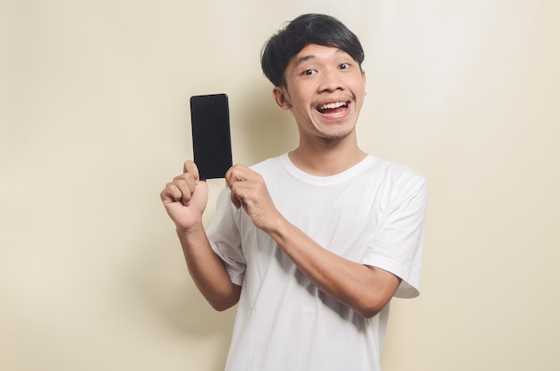 Feliz homem asiático vestindo camiseta branca brilhante com gesto mostrando a tela do telefone em fundo isolado