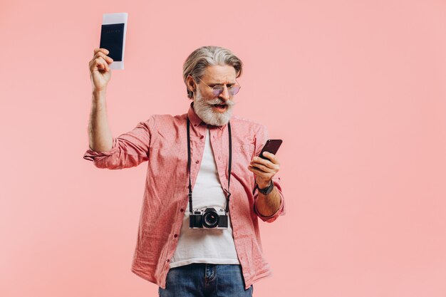 Feliz homem barbudo de óculos escuros, segurando um telefone celular e um passaporte com bilhetes na rosa