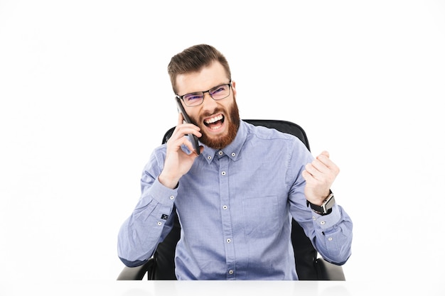 Feliz homem barbudo elegante em óculos falando por smartphone e se alegra sentado à mesa