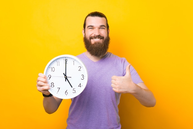 Feliz homem barbudo está segurando um relógio redondo e aparecendo o polegar.