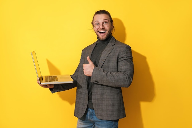 Feliz homem barbudo usando óculos está usando laptop e dando o sinal de aprovação do polegar comum