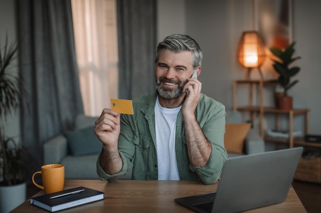 Feliz homem caucasiano aposentado com chamadas de barba por smartphone usa cartão de crédito e pc para fazer compras on-line