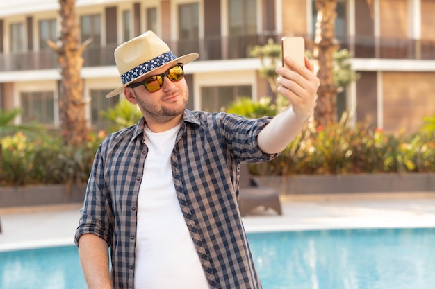 Feliz homem caucasiano com óculos de sol usando smartphone perto de hotel Turista de férias