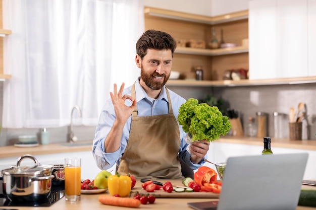 Feliz homem caucasiano do milênio cozinhando come salada e sinal de mão ok na mesa com legumes e computador