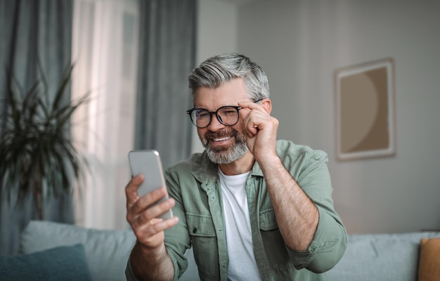 Feliz homem caucasiano sênior com barba em óculos olha para smartphone tem boas notícias no espaço de cópia interior da sala