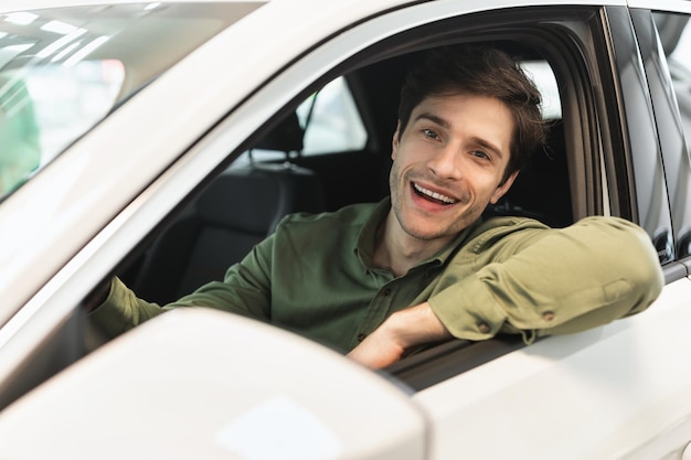 Feliz homem do milênio sentado no banco do motorista do novo automóvel após a compra olhando pela janela e