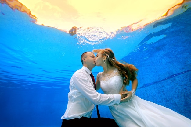 Foto feliz homem e mulher em vestidos de noiva beijando debaixo d'água na piscina no fundo de um pôr do sol