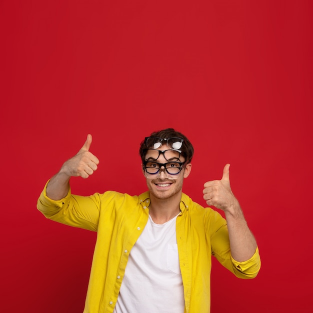 feliz homem engraçado na camisa amarela com muitos óculos no rosto aparecendo polegares isolado em fundo vermelho