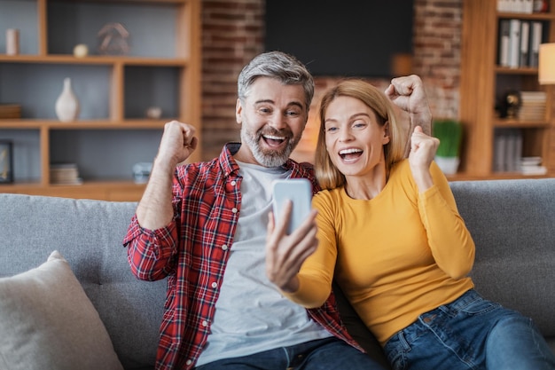 Feliz homem europeu adulto animado e mulher olhando para smartphone fazendo gesto de vitória comemorando vitória