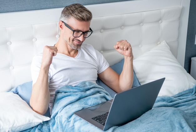 Feliz homem maduro em óculos trabalhando no laptop na cama