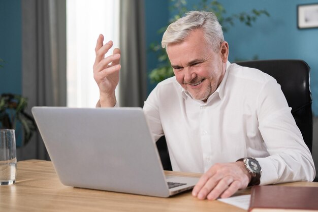 Feliz homem maduro sentado à mesa em casa olha para a tela do dispositivo usando o aplicativo de videochamada no laptop