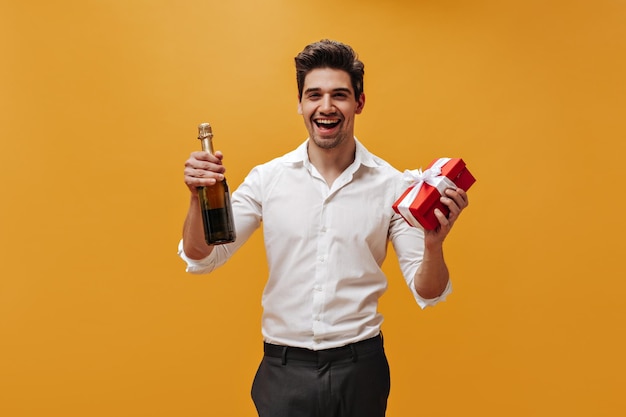 Feliz homem moreno alegre na camisa branca estilo clássico e sorrisos de calças pretas sinceramente segura garrafa de champanhe e caixa de presente vermelha em fundo laranja