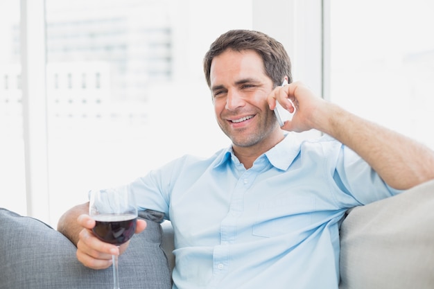 Feliz homem relaxando no sofá com um copo de vinho tinto falando no telefone