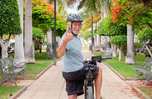 Feliz homem sênior ativo andando de bicicleta no parque público com bicicleta elétrica olhando para a câmera com o polegar para cima Conceito de estilo de vida saudável e mobilidade sustentável