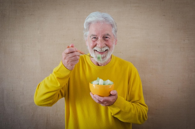 Feliz homem sênior de cabelos brancos na camisa amarela isolada em um fundo claro está segurando uma tigela com melão cortado pronto para ser comido