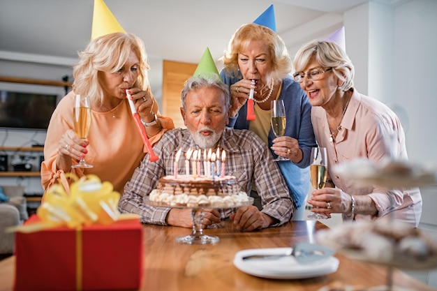 Feliz homem sênior soprando velas de aniversário em um bolo enquanto faz festa de aniversário com amigos em casa