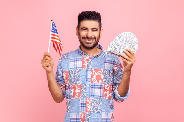 Feliz homem sorridente com barba, vestindo camisa azul estilo casual, olhando para a câmera segurando notas e bandeira dos EUA nas mãos, expressando emoções positivas Foto de estúdio interior isolada no fundo rosa