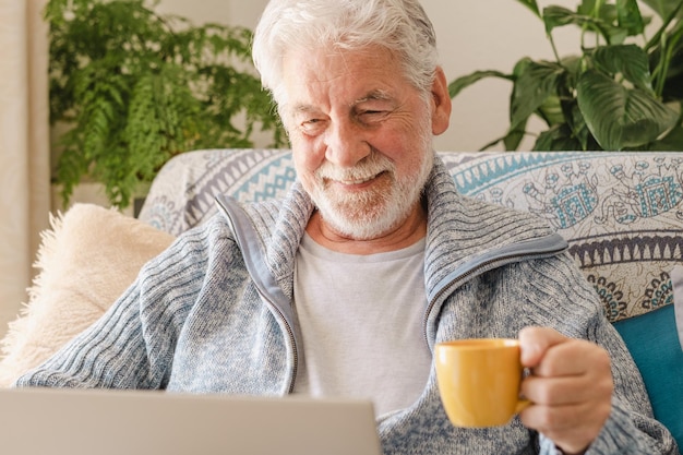 Feliz idoso dos anos 70 sentado no sofá navegando pelo laptop segurando uma xícara de café geração mais antiga e conceito de uso fácil de aplicativo de tecnologia moderna