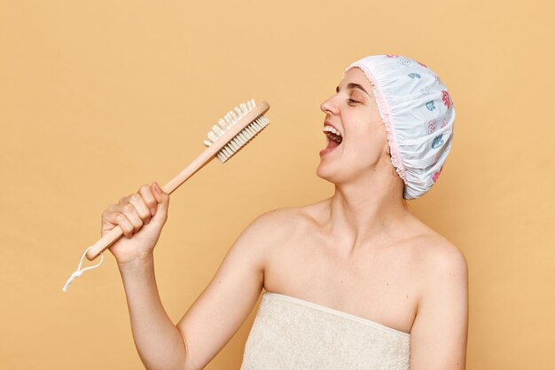 Feliz jovem adulta animada com escova de massagem fingindo seu microfone cantando se divertindo enquanto tomava banho em pé isolado no fundo bege