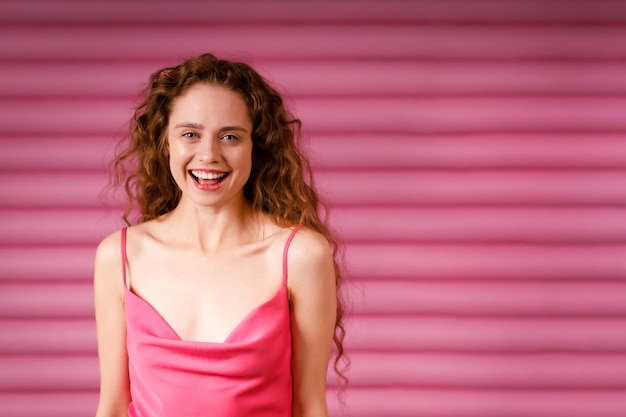 Feliz jovem adulta sorridente usando vestido rosa dentro de casa olhando para a câmera com um sorriso alegre em fundo rosa