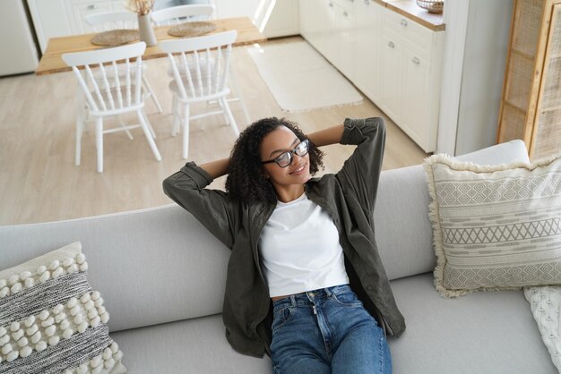 Feliz jovem afro-americana relaxando no sofá confortável na aconchegante sala de estar aproveitando o intervalo