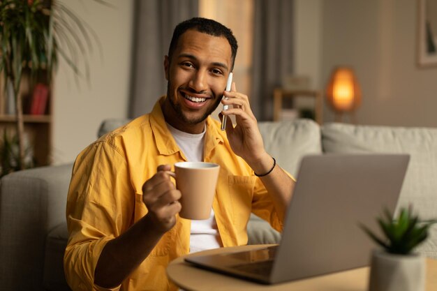 Feliz jovem afro-americano com café falando no celular usando laptop pc no escritório em casa