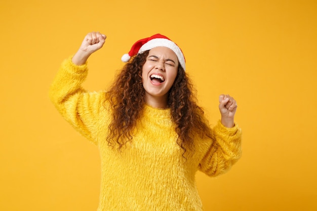 Feliz jovem afro-americano Papai Noel com chapéu de Natal isolado em fundo amarelo. Feliz ano novo 2020 conceito de feriado de celebração. Simule o espaço da cópia. Fazendo gesto de vencedor, mantendo os olhos fechados.