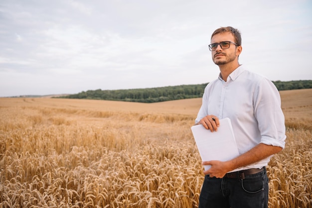 Feliz jovem agricultor engenheiro com o caderno de pé no campo de trigo enquanto colheitadeira trabalhando em segundo plano