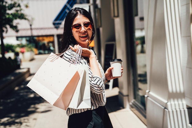 feliz jovem asiática alegremente rosto câmera rindo. mulher de óculos de sol segurando sacos de café compras ao ar livre na tomada do shopping center sob o sol. bela dama de pé na rua pela loja de tecidos