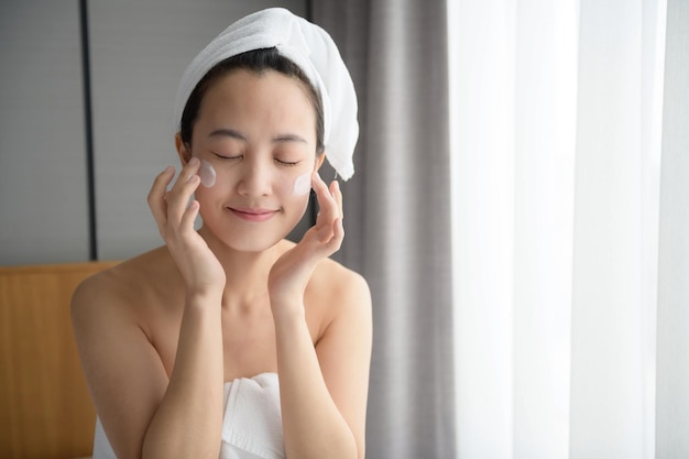 Feliz jovem asiática aplicando loções faciais enquanto usava uma toalha e tocando seu rosto Maquiagem diária e cuidados com a pele