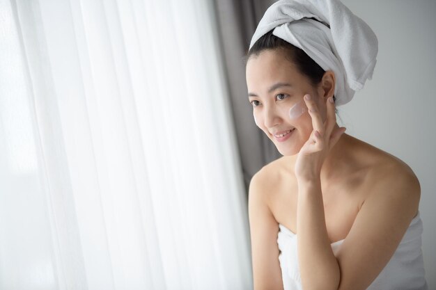 Feliz jovem asiática aplicando loções faciais enquanto usava uma toalha e tocando seu rosto Maquiagem diária e cuidados com a pele
