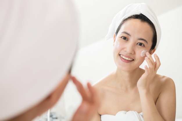 Feliz jovem asiática aplicando loções para o rosto enquanto usava uma toalha e tocando seu rosto no banheiro
