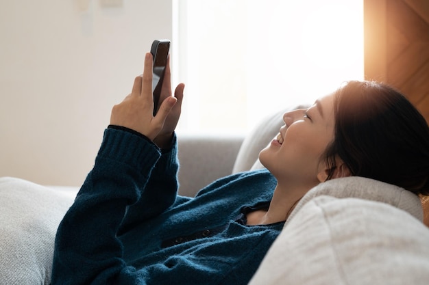 Feliz jovem asiática deitada no sofá na sala de estar e usando o celular em casa