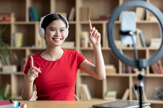 Feliz jovem asiática em fones de ouvido sem fio gesticulando com as mãos e olhe para a webcam do smartphone