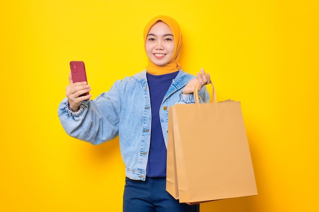 Feliz jovem asiática em jaqueta jeans segurando celular e sacolas de compras olhando para câmera isolada sobre fundo amarelo