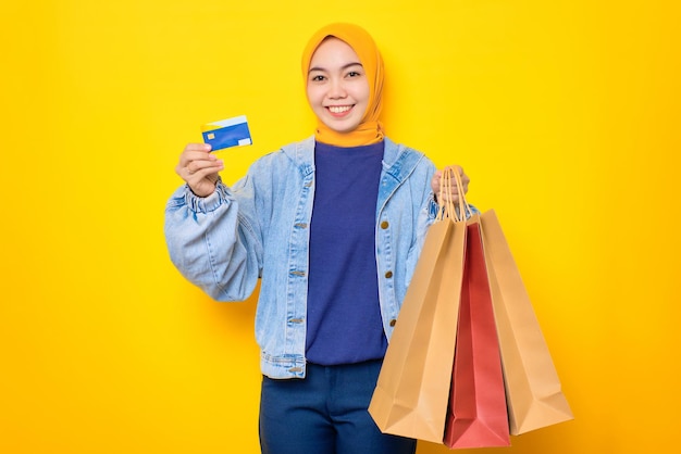 Feliz jovem asiática em jaqueta jeans segurando sacolas de compras e cartão de crédito olhando para câmera isolada sobre fundo amarelo