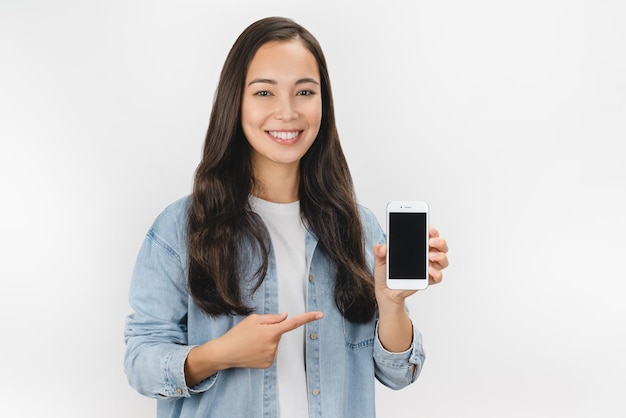 Feliz jovem asiática segurando o celular de tela em branco e apontando o dedo sobre fundo branco