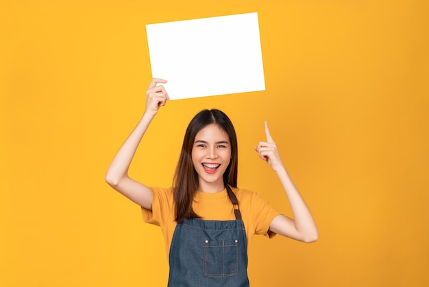 Feliz jovem asiática usa um avental e segurando um papel em branco com uma cara sorridente e olhando para o fundo laranja. para sinais de publicidade.