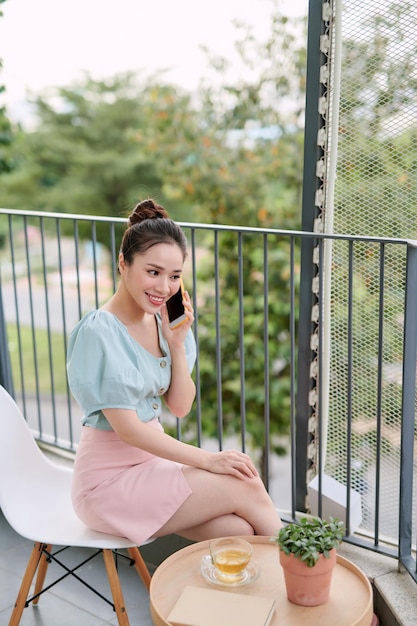 Feliz jovem asiática usando telefone quando sentada bebendo chá na varanda