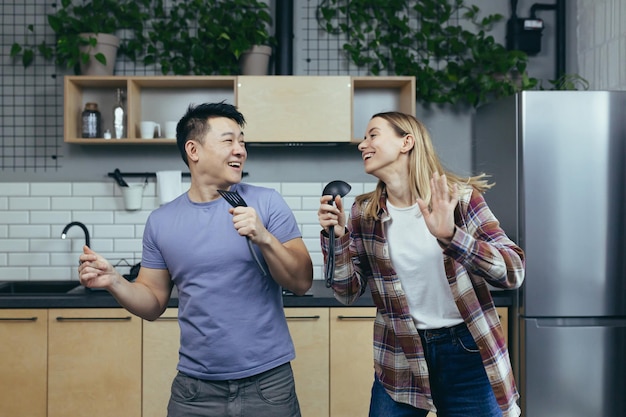 Feliz jovem asiático e casal de mulheres cantando e dançando juntos na cozinha se divertindo juntos família apaixonada