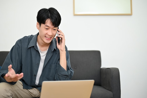 Feliz jovem asiático olhando para a tela do laptop falando ao telefone com seus colegas