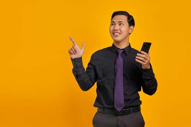 Feliz jovem asiático segurando um telefone celular e apontando o dedo para copiar o espaço no fundo amarelo