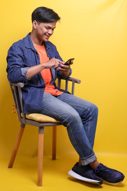 Feliz jovem asiático sentado relaxando na cadeira e usando smartphone sobre fundo amarelo