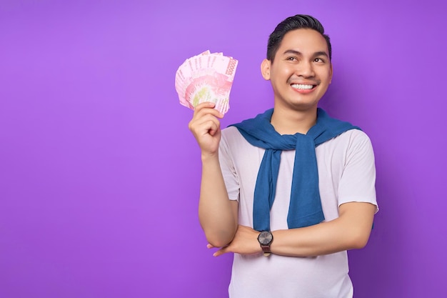 Feliz jovem asiático vestindo camiseta branca roupas casuais segurando notas de dinheiro e sorrindo de lado isolado sobre fundo roxo Conceito de lucro e riqueza