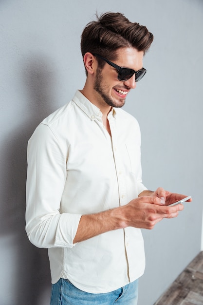 Feliz jovem atraente usando óculos escuros em pé e usando o smartphone