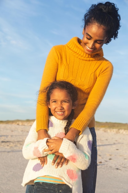 Feliz jovem birracial abraçando a filha por trás em pé na praia contra o céu