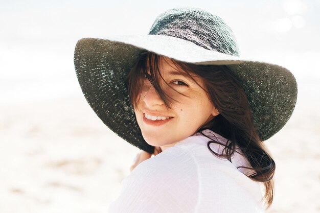 Feliz jovem boho de chapéu relaxando e aproveitando o dia ensolarado e quente no oceano Espaço para texto Menina elegante hipster sentada na praia e bronzeando Férias de verão Emoções calmas