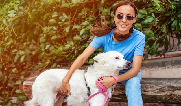 Feliz jovem bonita garota morena abraçando seu cão fofo branco bonito ao ar livre no parque no gramado adoção resgatado abrigo companheiro animal de estimação